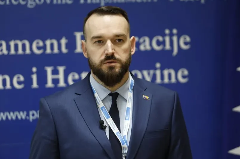 Muškarac u tamnoplavom odijelu i tamnoj kravati, sa zlatnom značkom Bosne i Hercegovine na reveru, stoji ispred plave pozadine sa bijelim natpisom.