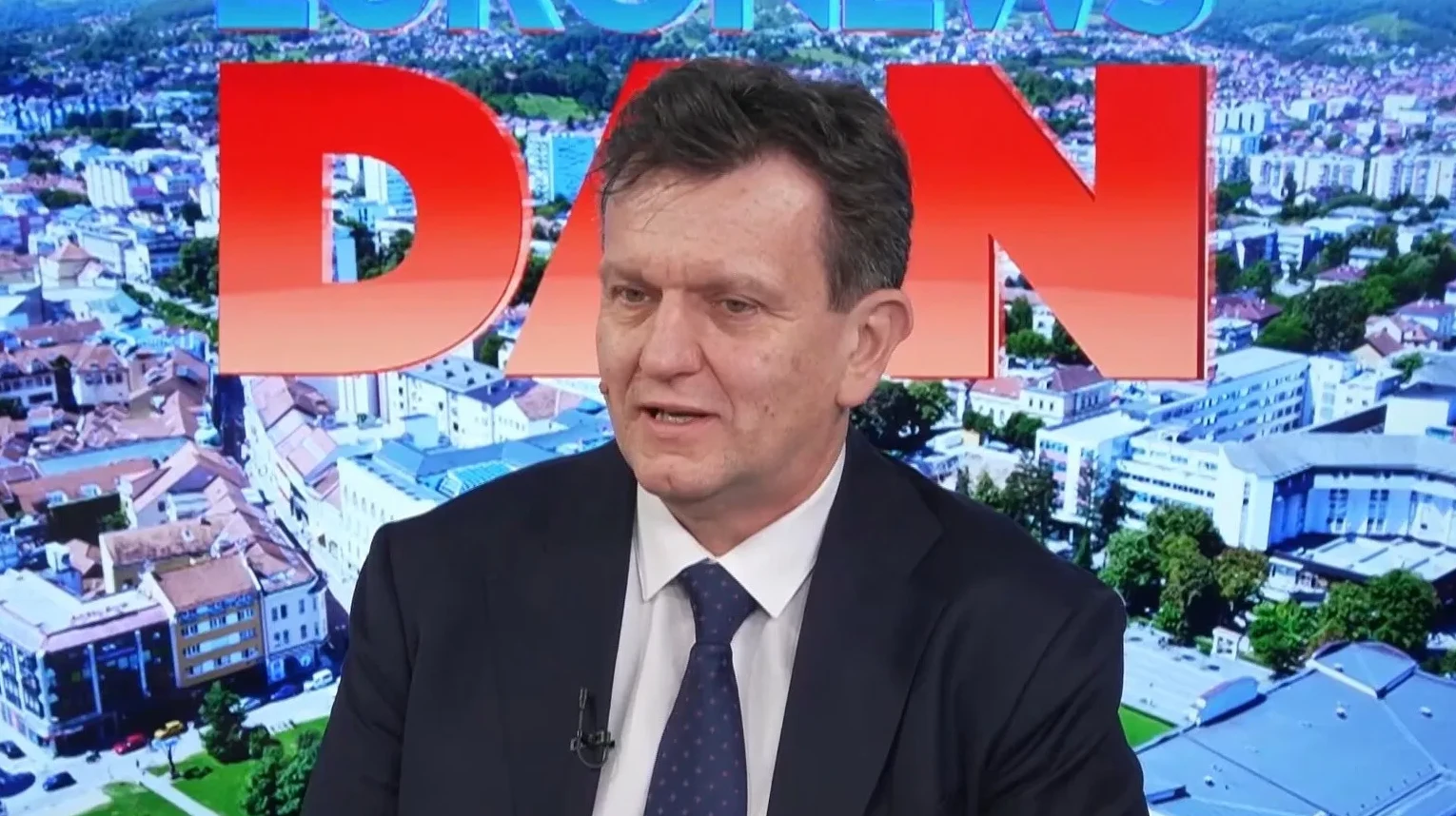 Političar u tamnom odijelu i plavoj kravati s bijelom košuljom, sjedi u studiju i govori pred kamerom. U pozadini je grafika s gradom i velikim crvenim slovima 'DAN' i logom 'EURONEWS'.