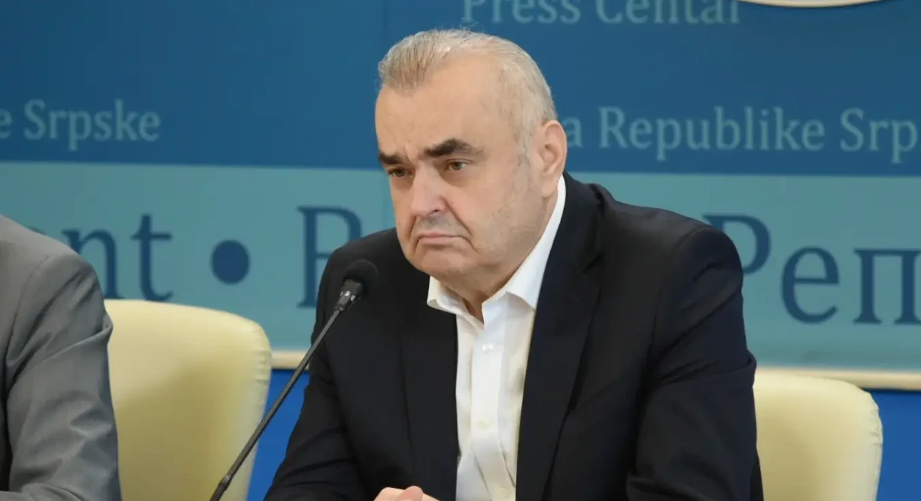 Portret starijeg gospodina u crnom odijelu i bijeloj košulji, kako sjedi za govornicom ispred plave pozadine s natpisom.