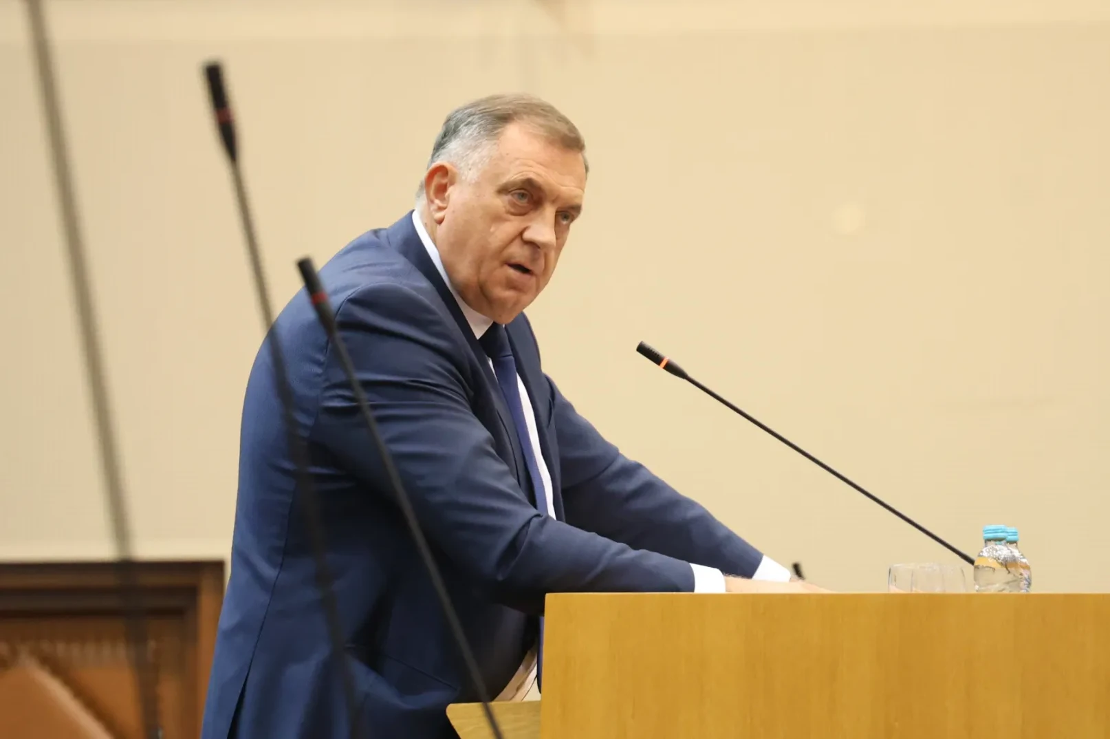 Političar u tamnom odijelu govori za govornicom, dok se na stolu ispred njega nalazi boca vode.