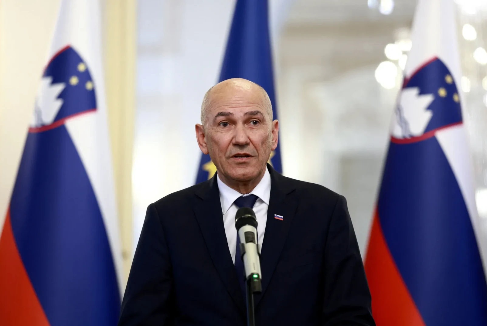 Političar u tamnom odijelu i plavoj kravati, s predsjedničkim znakom sa slovenskom zastavom, stoji pred mikrofonom, a iza njega su istaknute tri zastave Slovenije.