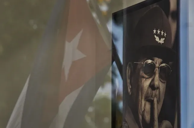 Crno-bijeli portret Fidela Castra sa podignutim prstom na usnama, uz odraz kubanske zastave.