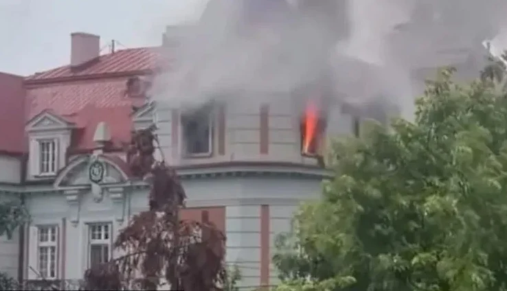 Vatra izbija iz prozora zgrade okružene dimom, sa crvenim krovom i drvećem u prednjem planu.