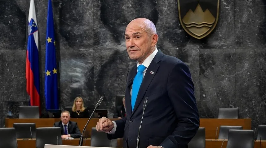 Političar u tamnom odijelu i plavoj kravati obraća se publici u skupštinskoj sali, sa zastavom Slovenije i Evropske unije iza njega.