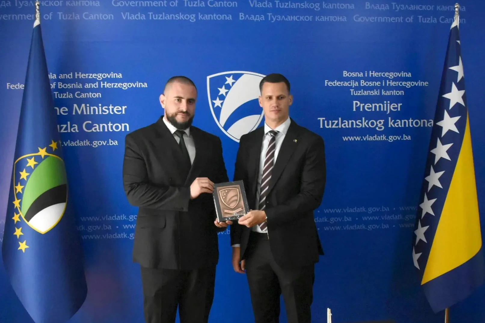 Dva zvaničnika, premijer i ministar Tuzlanskog kantona, poziraju za fotografiju u službenim odijelima pored zastava Bosne i Hercegovine i Tuzlanskog kantona.