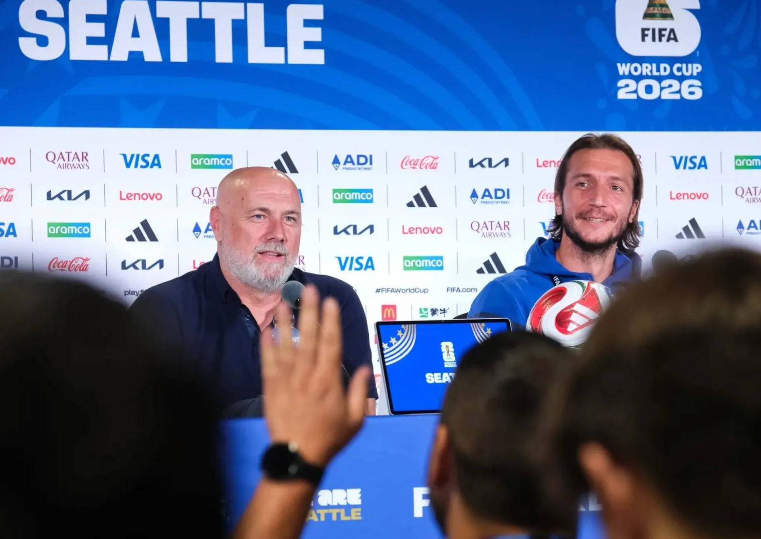 Dva muškarca na pres-konferenciji, jedan s loptom u ruci, sa natpisom "FIFA WORLD CUP 2026 SEATTLE"