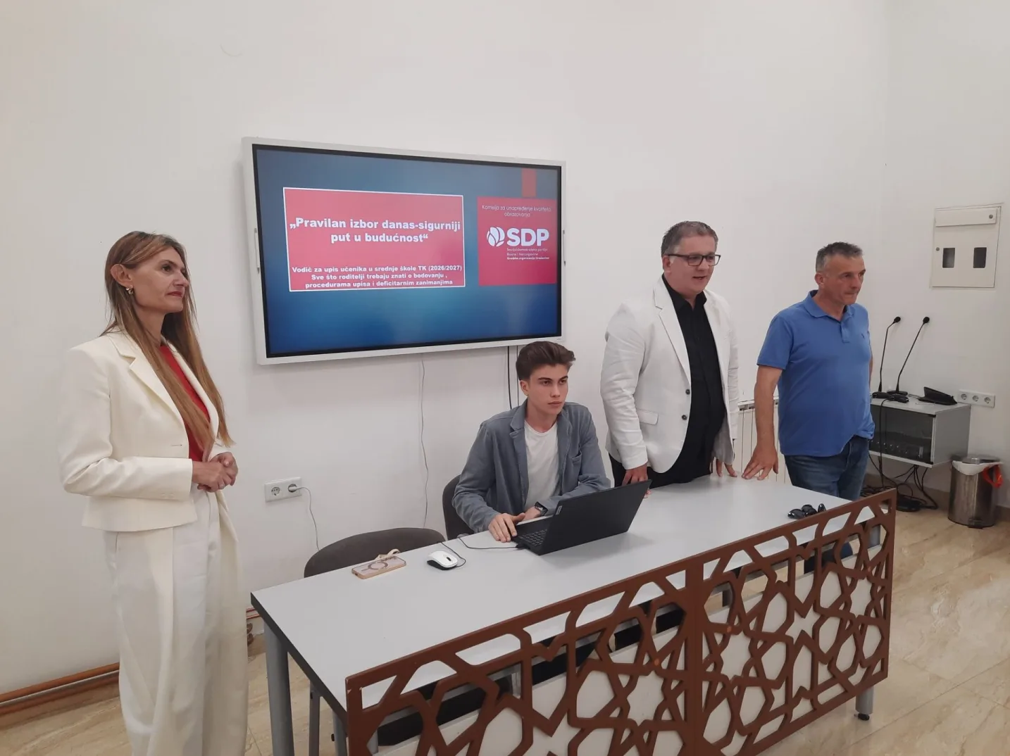 Četiri osobe stoje oko stola ispred ekrana sa prezentacijom SDP-a.