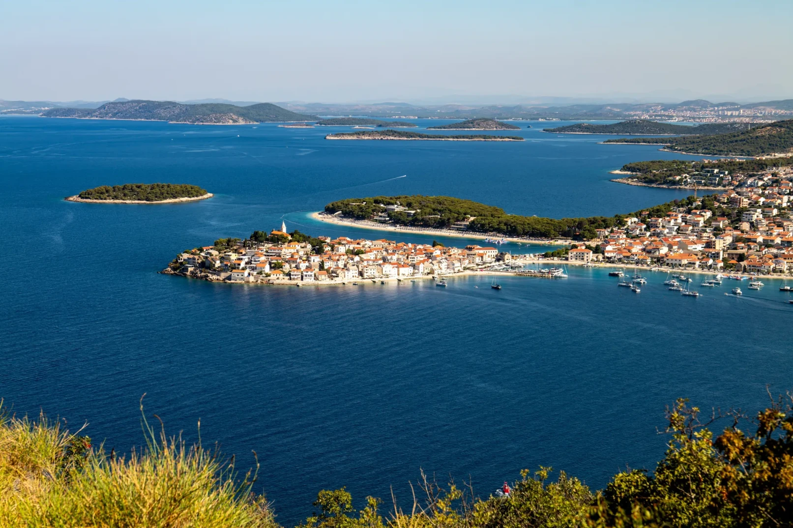Pogled na primorski grad sa ostrvom okruženim plavim Jadranskim morem, sa brojnim brodovima usidrenim u luci.