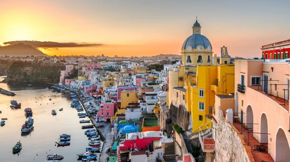 Pogled na živopisni mediteranski zaliv Procida sa šarenim kućama, crkvom sa kupolom i brojnim brodićima usidrenim u luci, osvijetljenim toplim zracima zalazećeg sunca.