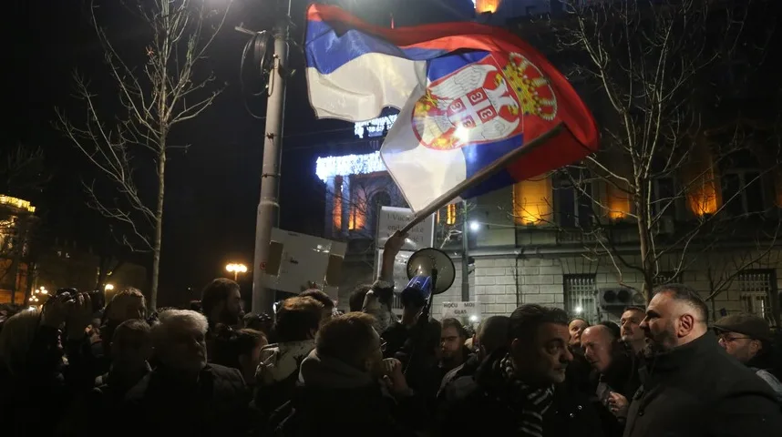 Grupa ljudi na noćnom protestu u Srbiji, sa zastavom Srbije koja se vijori.