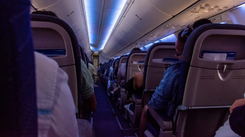 Unutrašnjost aviona sa putnicima koji sjede na svojim sjedištima tokom leta, osvijetljena plavičastom ambijentalnom rasvjetom.