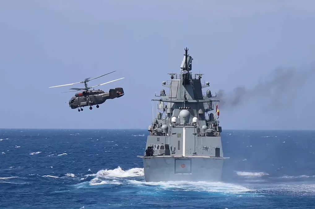 Ratni brod plovi po nemirnom moru, dok iznad njega leti vojni helikopter.