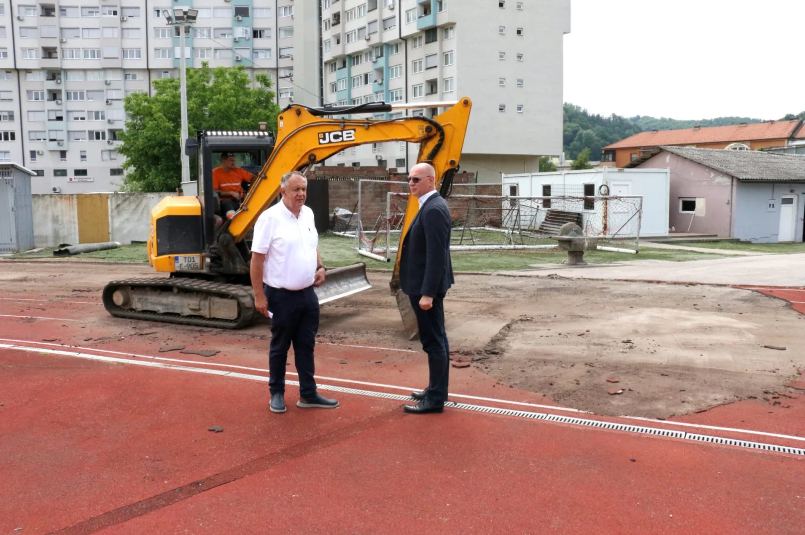 Dva muškarca stoje na crvenoj atletskoj stazi, a iza njih se vidi bager i gradilište.