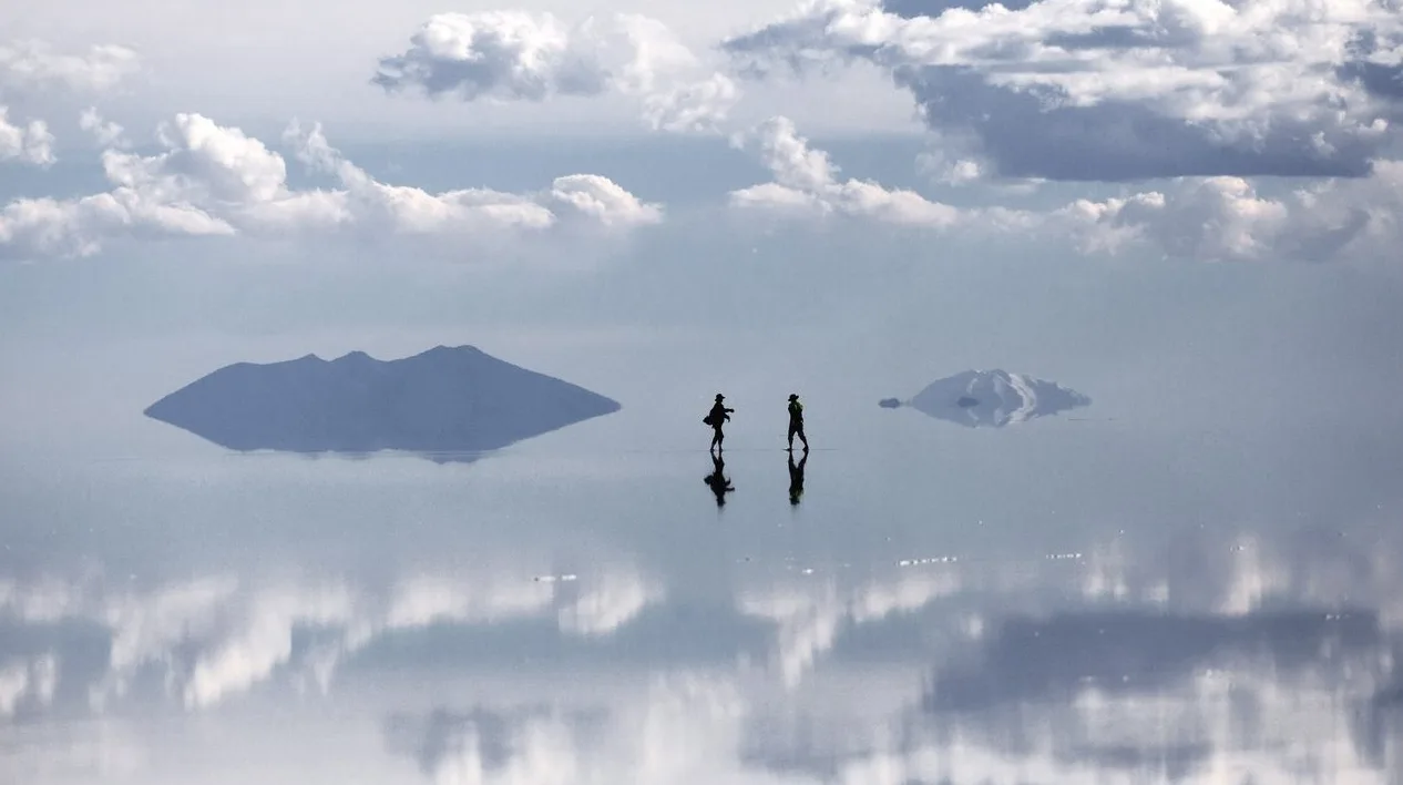 Dvoje ljudi šeta po zrcalnoj površini Salar de Uyuni, Bolivija, sa planinskim ostrvima koja se reflektuju u vodi.