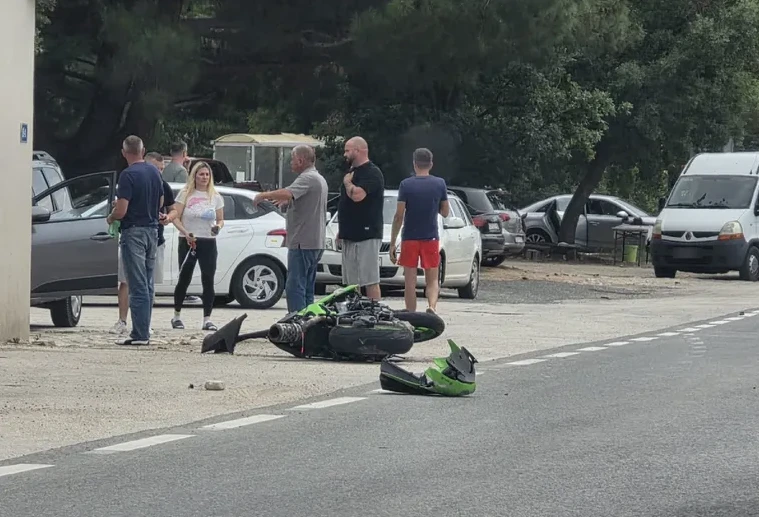 Policajci i građani okupljeni oko prevrnutog zelenog motocikla na putu, pored nekoliko automobila.