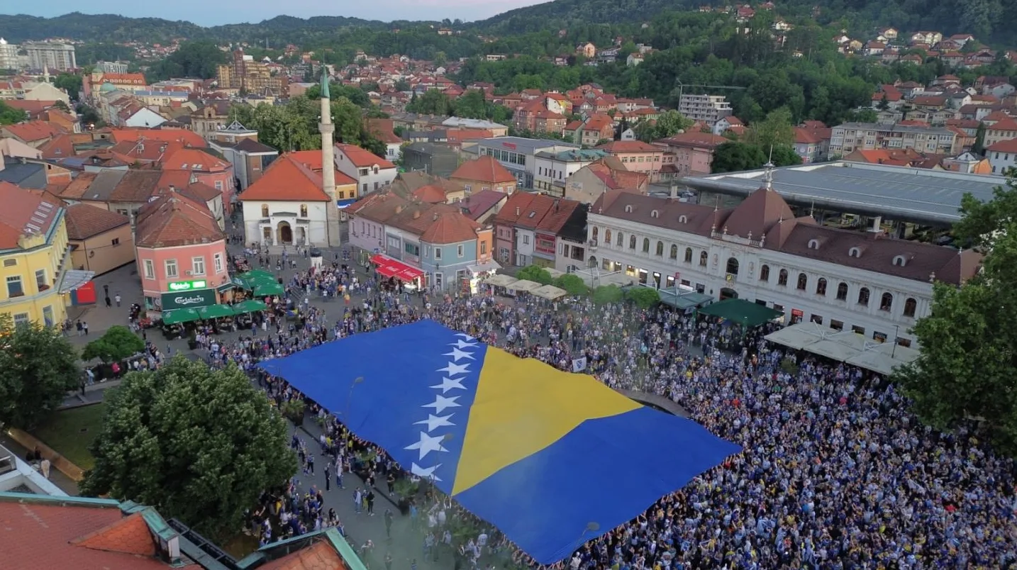 Velika zastava Bosne i Hercegovine razvijena na centralnom gradskom trgu okruženom mnoštvom ljudi i urbanom arhitekturom.
