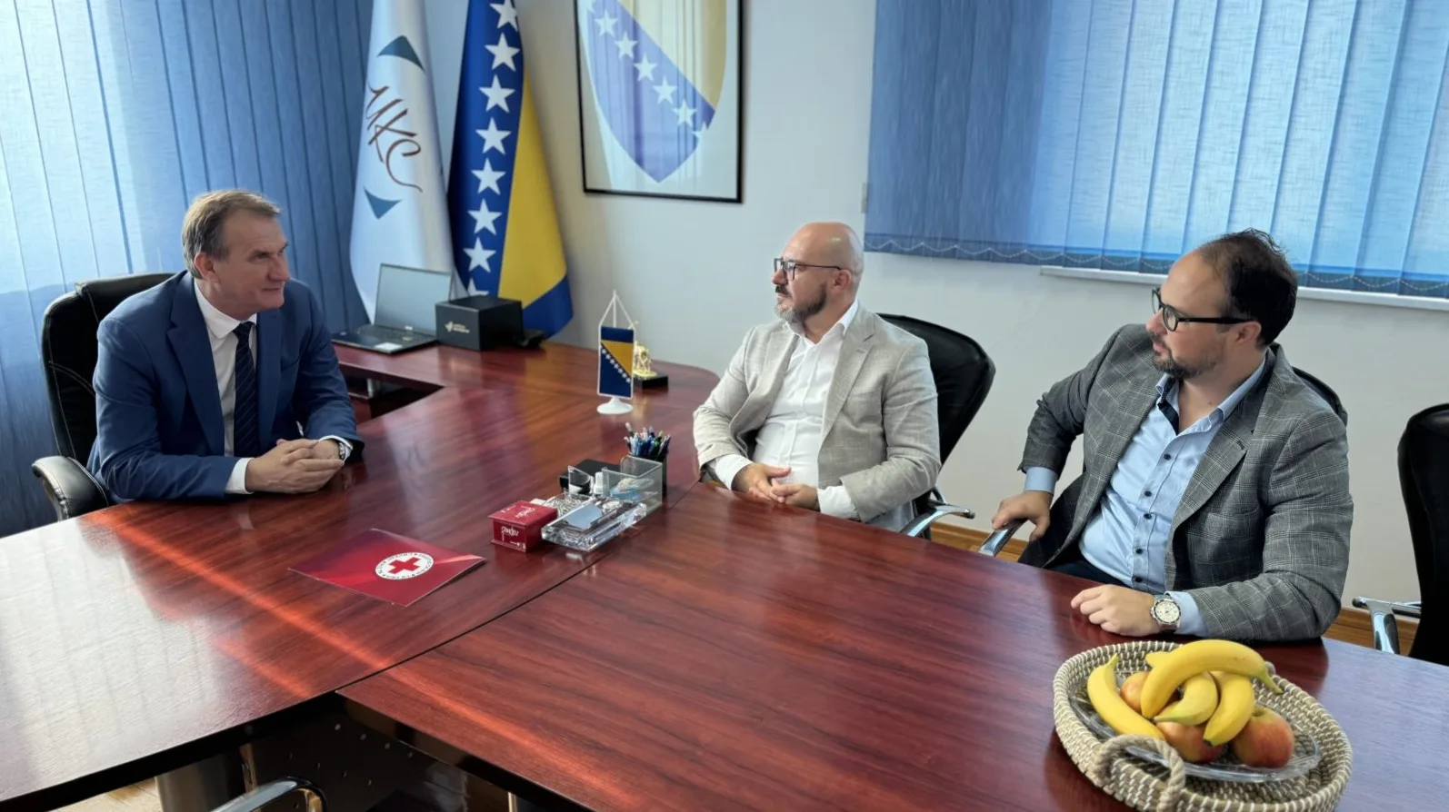 Troje muškaraca u odijelima sjede za velikim drvenim stolom u kancelariji, razgovarajući. Na stolu su zastave Bosne i Hercegovine, ukrasne figurice, olovke, cigarete i voće. Takođe se vidi logo Crvenog krsta.