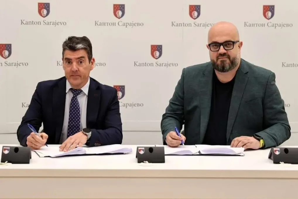 Dva muškarca u odijelu sjede za stolom i potpisuju dokumente u prostoriji sa grbom Kantona Sarajevo u pozadini.