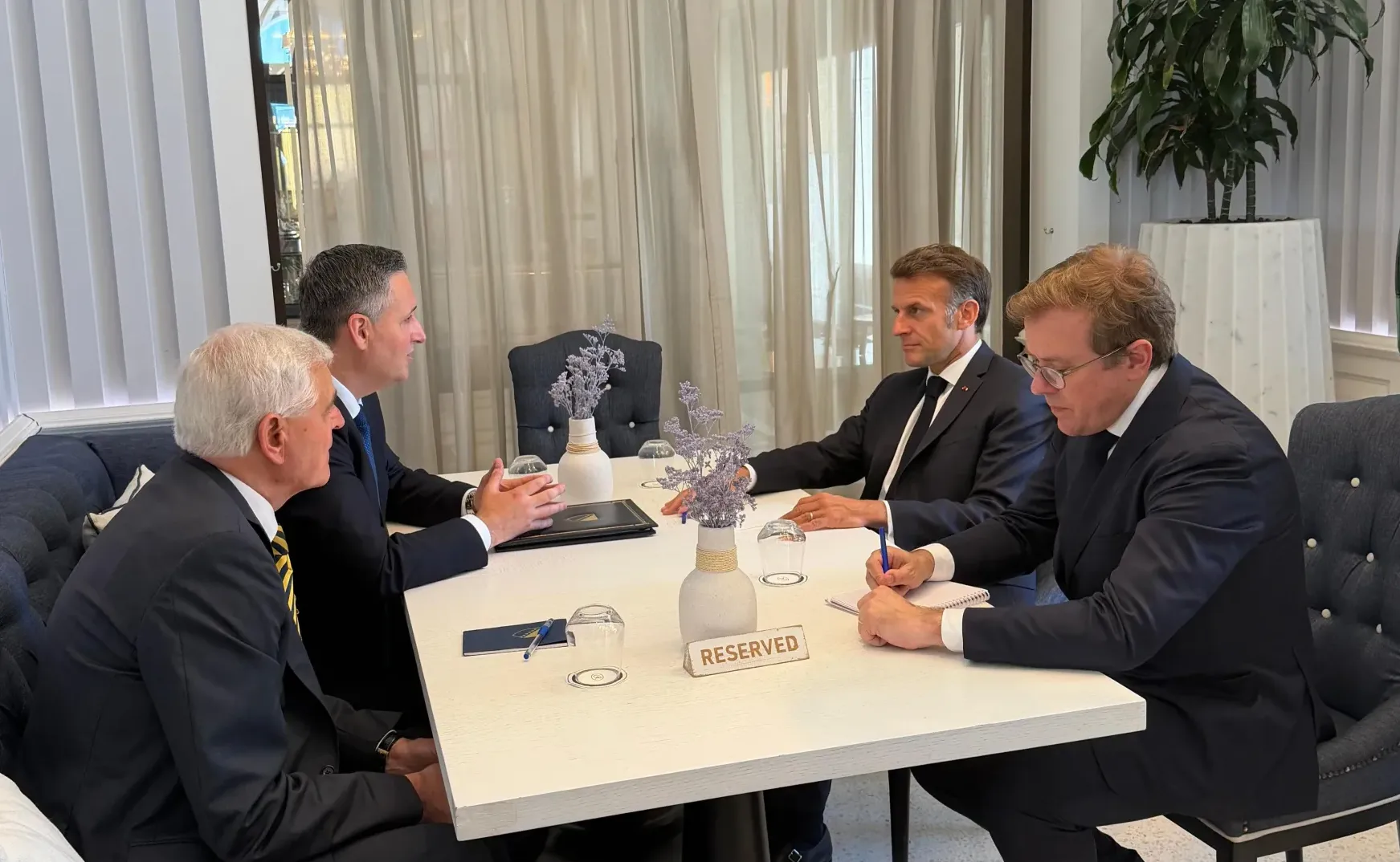 Četiri muškarca u odijelima sjede za bijelim stolom u restoranu, s natpisom "RESERVED" na stolu. Francuski predsjednik Emmanuel Macron sjedi preko puta dvojice muškaraca, dok treći piše nešto u bilježnicu.