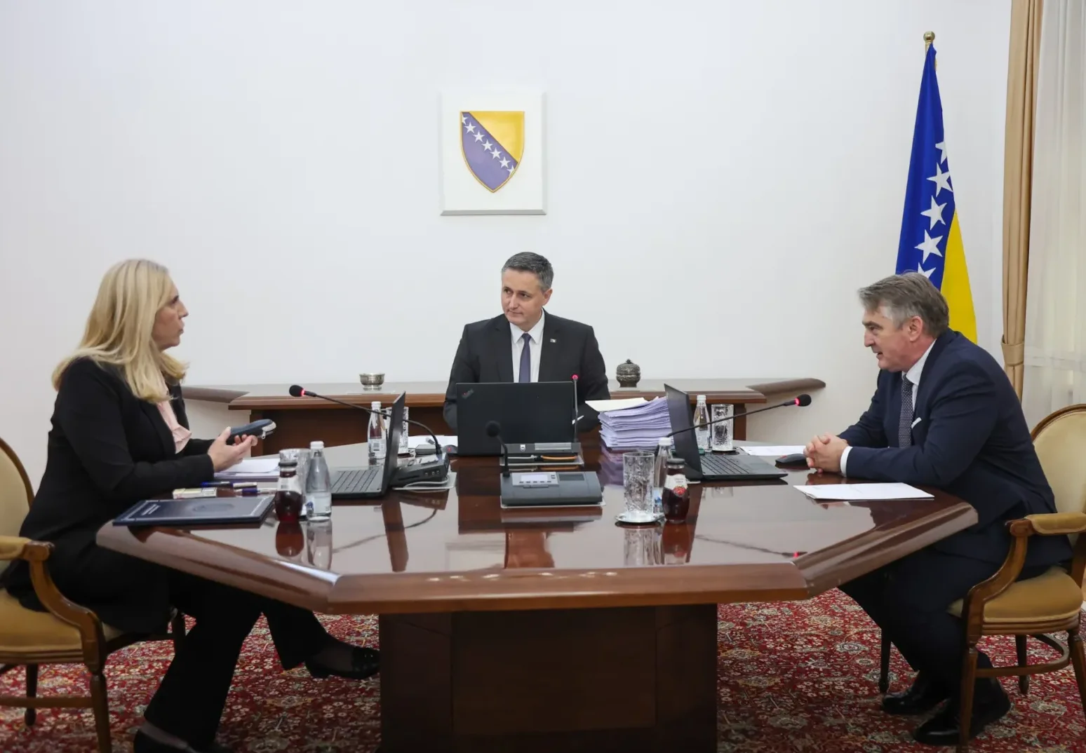 Tri osobe sjede za velikim drvenim stolom na sastanku, uz zastavu Bosne i Hercegovine u pozadini.