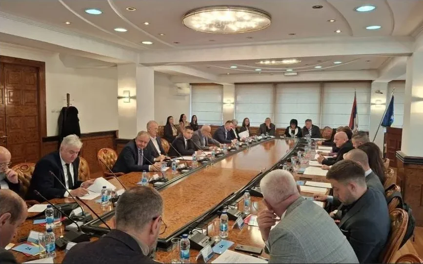 Grupa ljudi sjedi za dugačkim drvenim stolom na sastanku, ispred svakog sjedišta su papiri i boce s vodom.