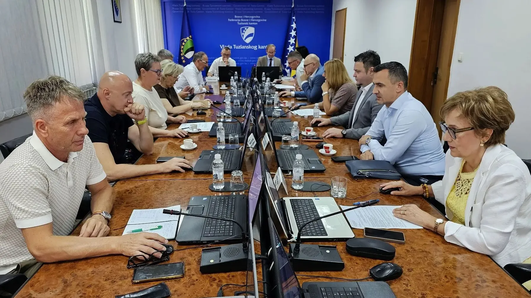 Grupa ljudi sjedi za dugim stolom u sali za sastanke, sa laptopima i dokumentima ispred sebe.