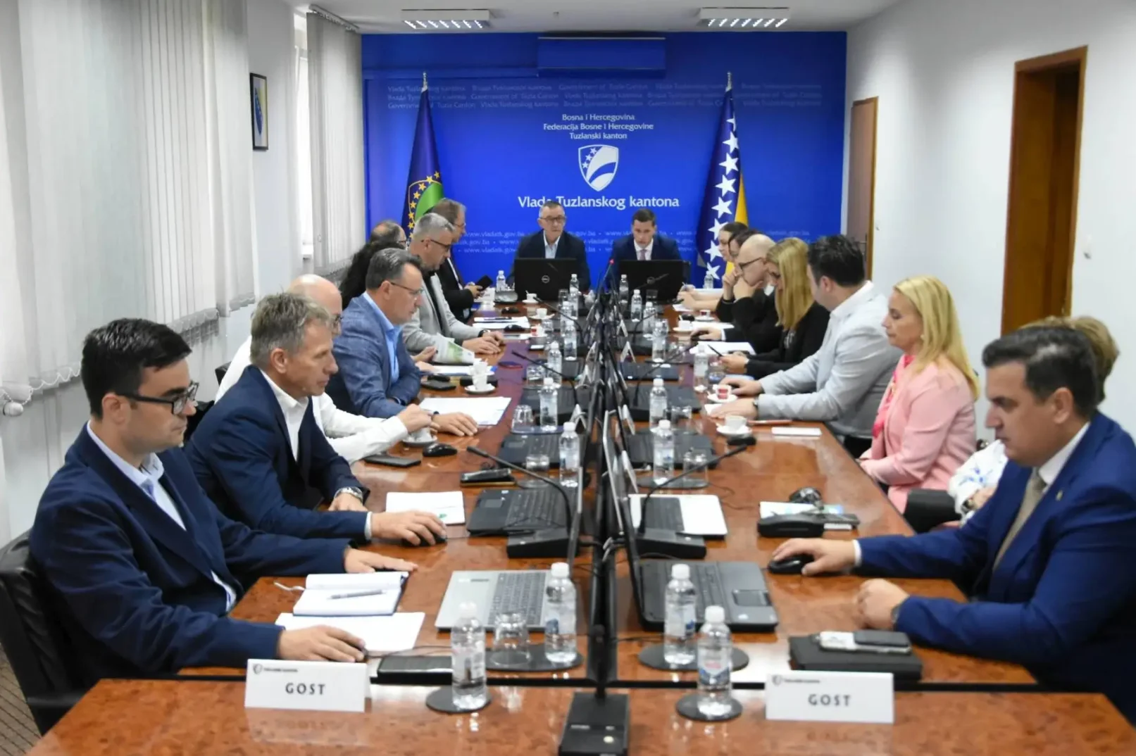 Grupa ljudi sjedi za dugačkim stolom u sali za sastanke, fokusirana na predmete ispred sebe. Na stolu su laptopi, bilježnice i boce s vodom.