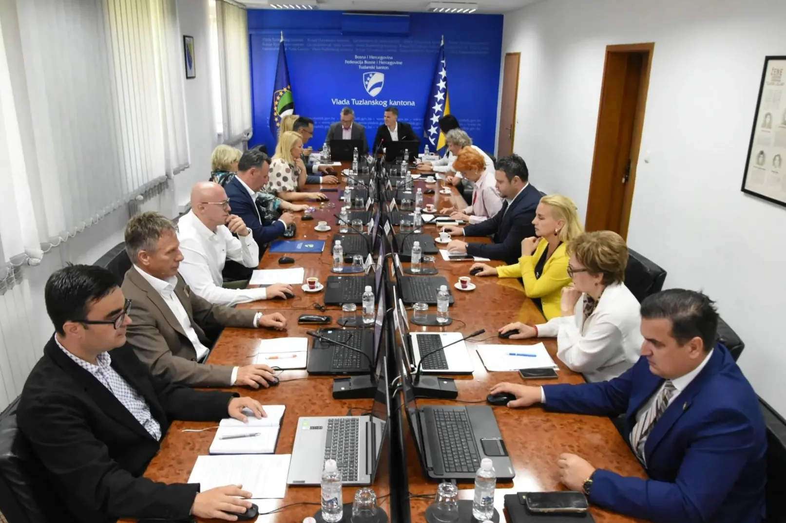 Grupa ljudi okupljena oko velikog drvenog stola na sastanku, svi sjede i gledaju prema naprijed, mnogi drže miševe od računara.