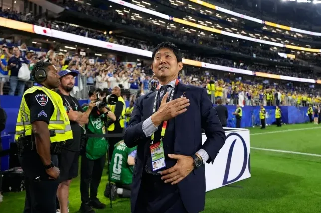 Fudbalski trener Japana sa rukom na srcu na stadionu, okružen publikom i obezbjeđenjem.