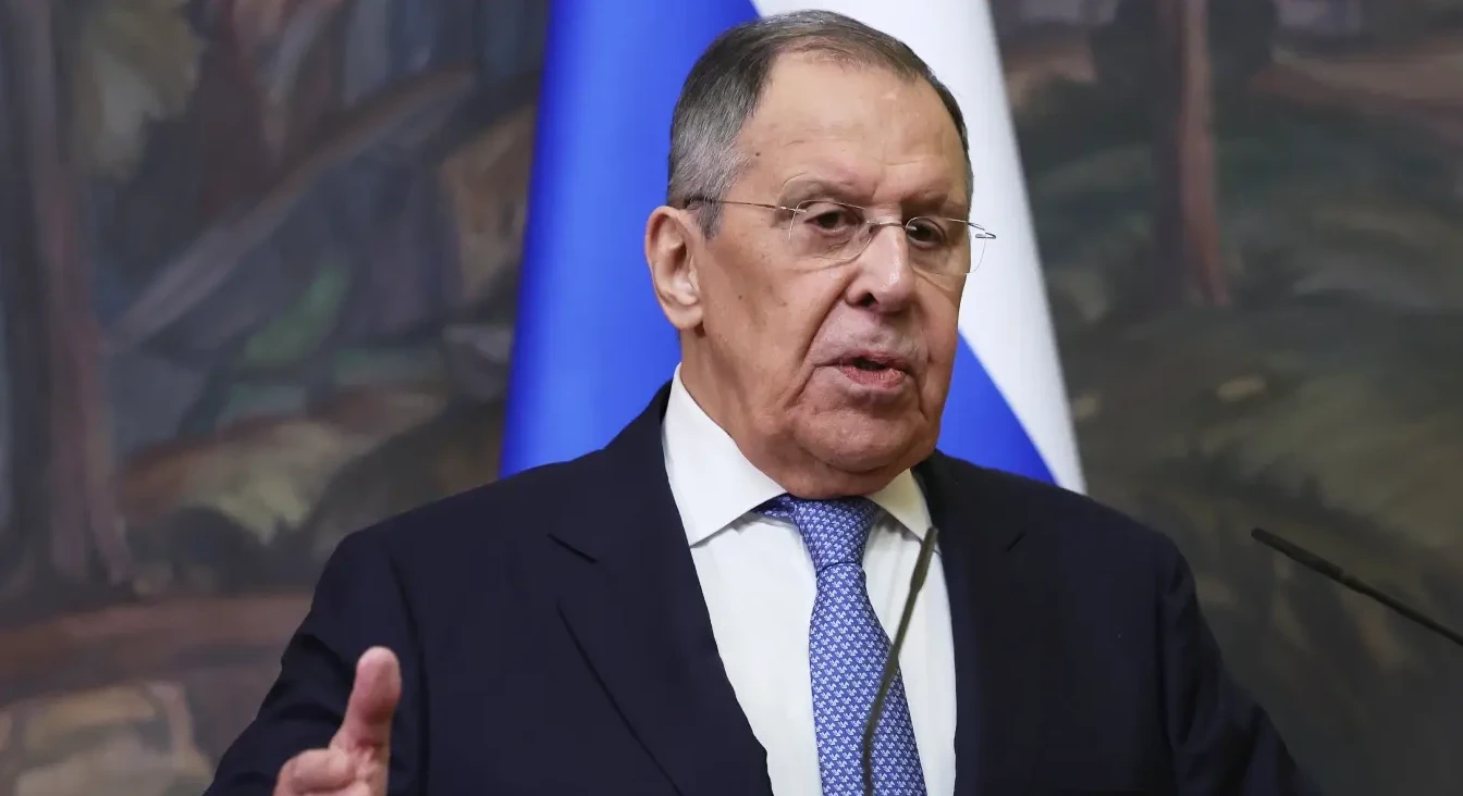 Sergej Lavrov nosi tamno odijelo, bijelu košulju i plavu kravatu sa dezenom, te naočare, dok govori za govornicom ispred ruske zastave.