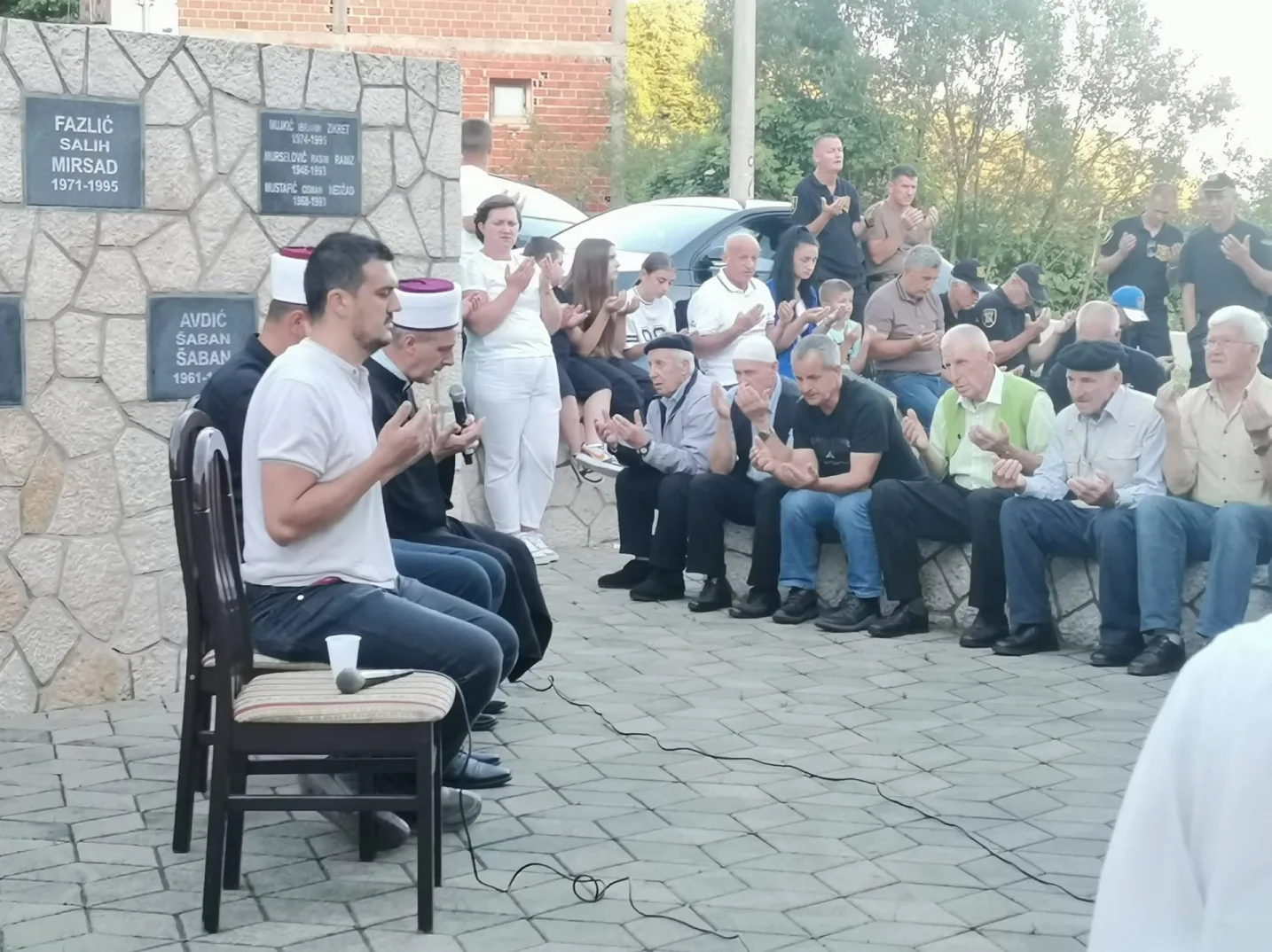 Grupa ljudi, većinom muškarci, okupljena ispred spomen-zida s uklesanim imenima i godinama. Mnogi od njih drže ruke sklopljene u molitvi, dok dvojica nose tradicionalne islamske kape. U prvom planu, na stolici pored mikrofona, sjedi muškarac u bijeloj majici. Atmosfera je svečana i sabrana.