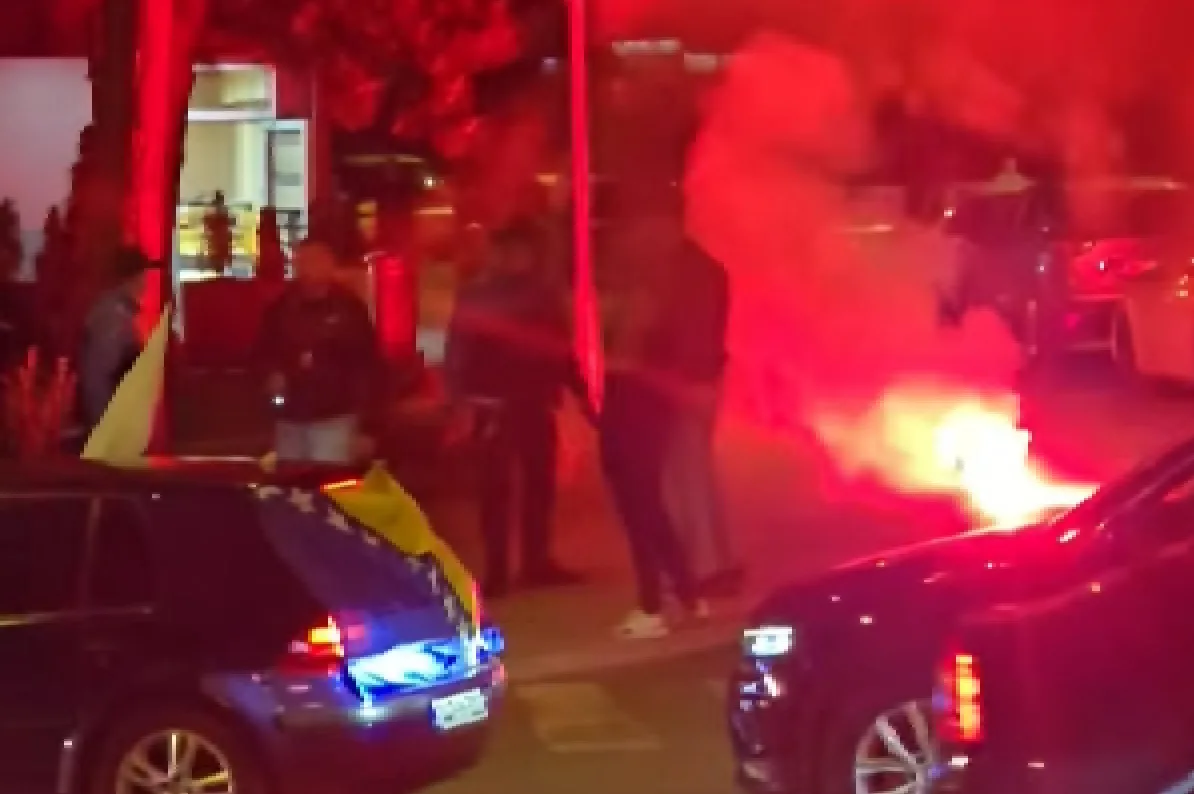 Skupina ljudi stoji na ulici u mraku, osvijetljena crvenim dimom iz bengalki i odsjajima svjetala automobila. Na automobilu u prvom planu vidi se dio zastave, a iza njega nazire se još jedna zastava.