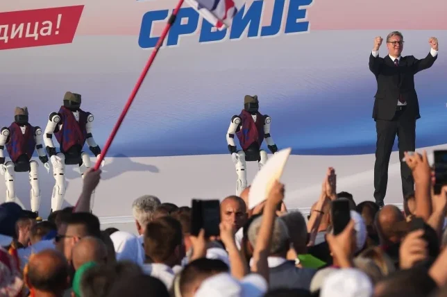 Čovjek u crnom odijelu diže ruke u zrak dok iza njega stoje roboti obučeni u narodne nošnje, a u pozadini se vidi natpis 'SRBIJE'.