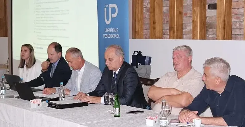 Učesnici sastanka Udruženja poslodavaca sjede za stolom ispred projekcionog platna i banera sa logom Udruženja.