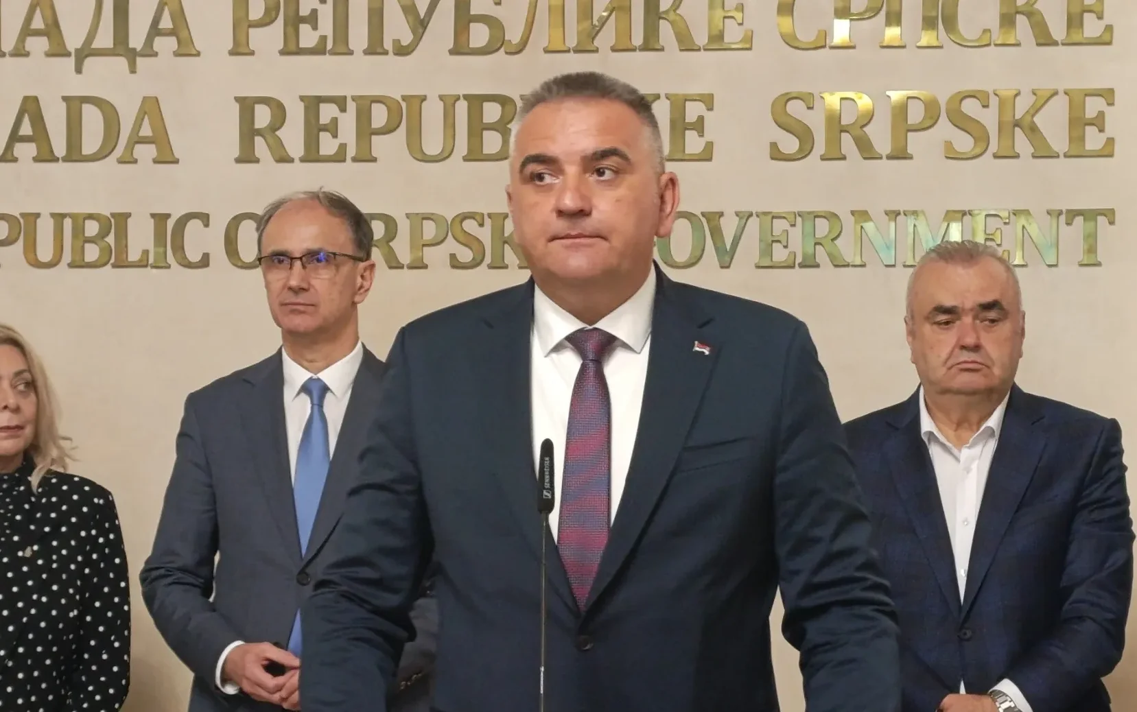 Muškarac u tamnom odijelu i crveno-plavoj kravati govori za govornicom, sa zastavom na reveru.