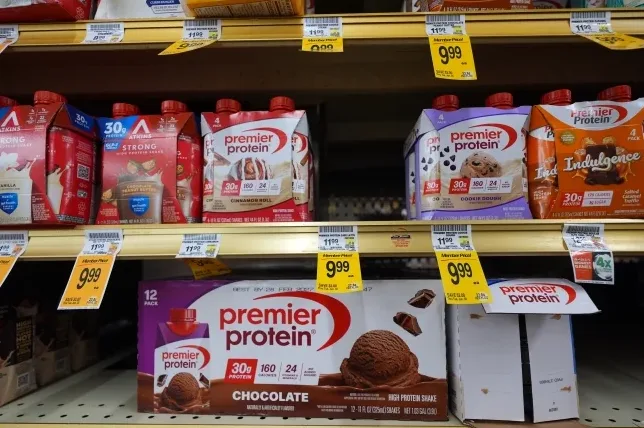 Različiti okusi proteinskih napitaka Premier Protein i Atkins na policama supermarketa, s cijenama istaknutim na žutim cjenovnim naljepnicama.
