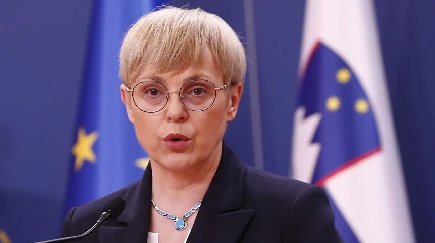 Političarka iz Slovenije sa naočarima i ogrlicom sa plavim kamenjem, govori ispred zastava Slovenije i Evropske Unije.