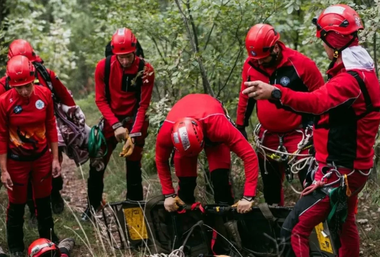 Grupa spasilaca u crvenim uniformama i kacigama radi u šumi, pripremajući nosila za transport.
