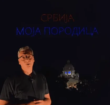 Čovjek govori ispred zgrade sa kupolom osvijetljenom plavom bojom, a iznad njih se u mraku ispisuju riječi "SRBIJA MOJA PORODICA" svijetlećim slovima.