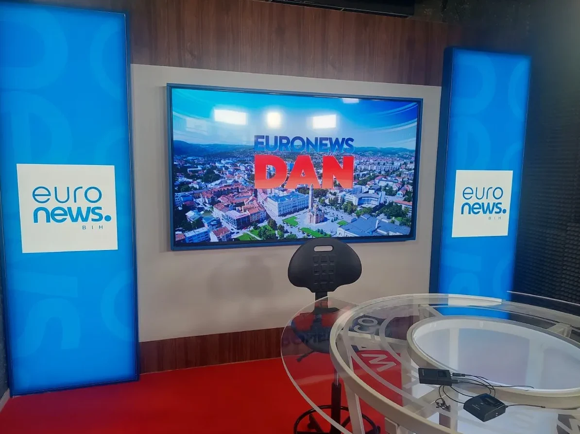 Unutrašnjost studija Euronews BiH s velikim ekranom na kojem piše "Euronews DAN", plavim osvijetljenim stupovima sa logom Euronews BiH i modernim stolom sa mikrofonima.