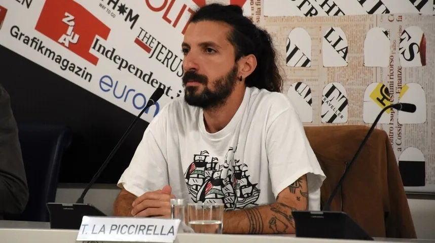 Čovjek s dugom kosom i bradom, obučen u bijelu majicu s printom jedrenjaka, sjedi za stolom ispred mikrofona i pozadine s logotipima medija.