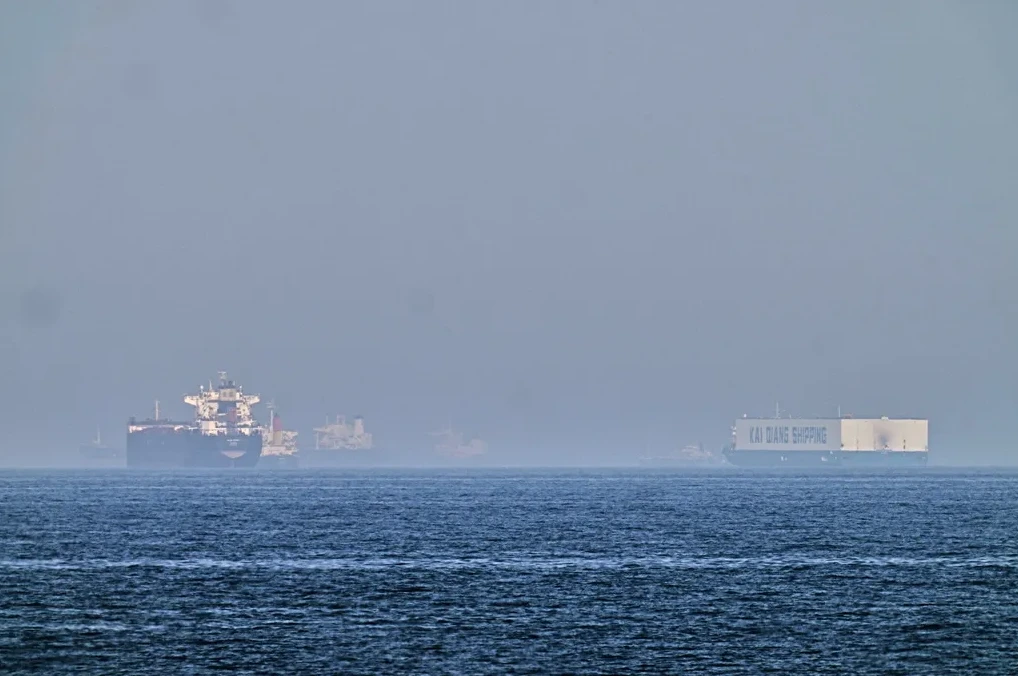 Kontejnerski brod sa natpisom "KAI QIANG SHIPPING" i drugi teretni brodovi na horizontu, na mirnom moru.