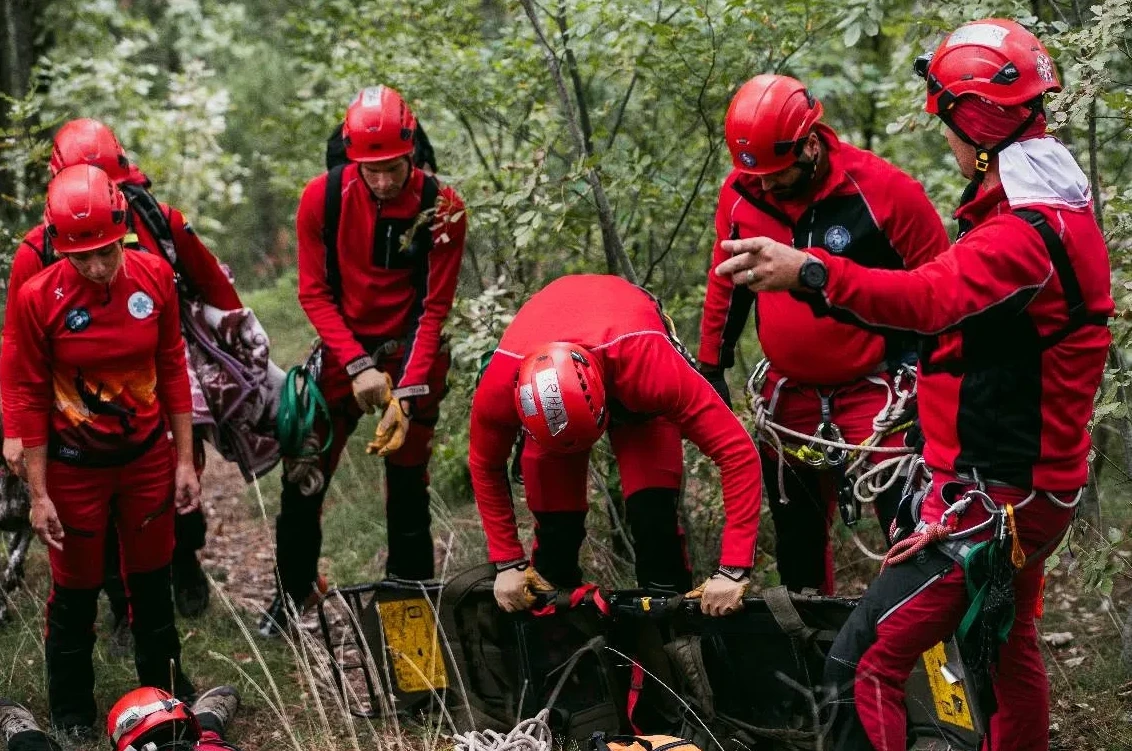 Članovi gorske službe spasavanja u crvenim uniformama i kacigama vježbaju transport povrijeđene osobe na nosilima u šumskom okruženju.