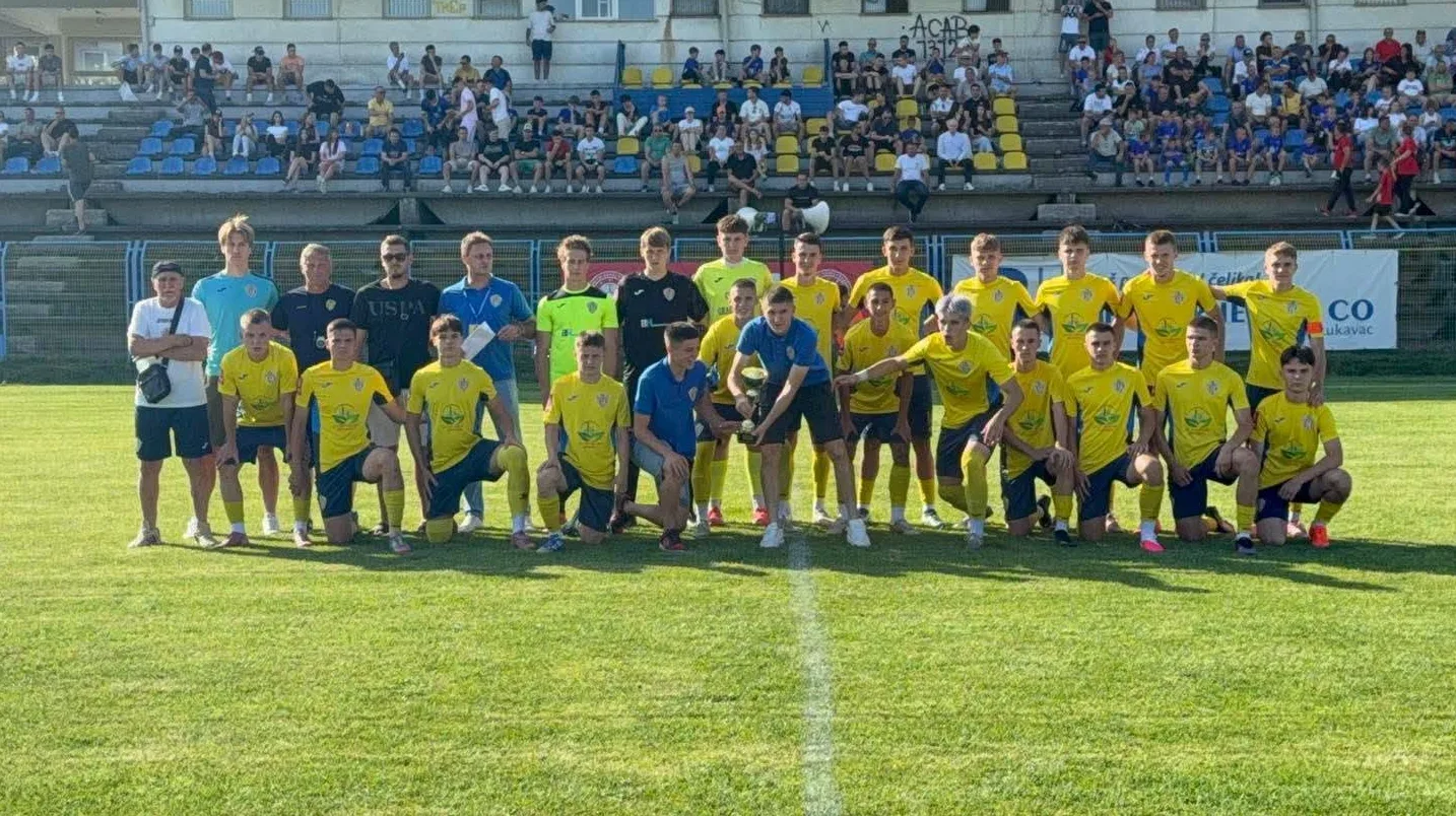 Fudbalski tim pozira za fotografiju na zelenom travnjaku, sa publikom na tribinama u pozadini.