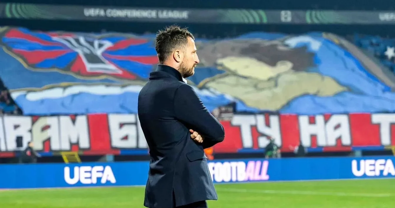 Trener u tamnom odijelu stoji na fudbalskom terenu sa rukama ukrštenim na prsima, gledajući u pravcu tribina iza sebe gdje se vide transparenti i navijači.