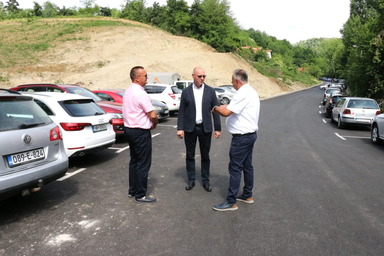 Tri muškarca u odijelima razgovaraju na novom parkingu pored niza parkiranih automobila.