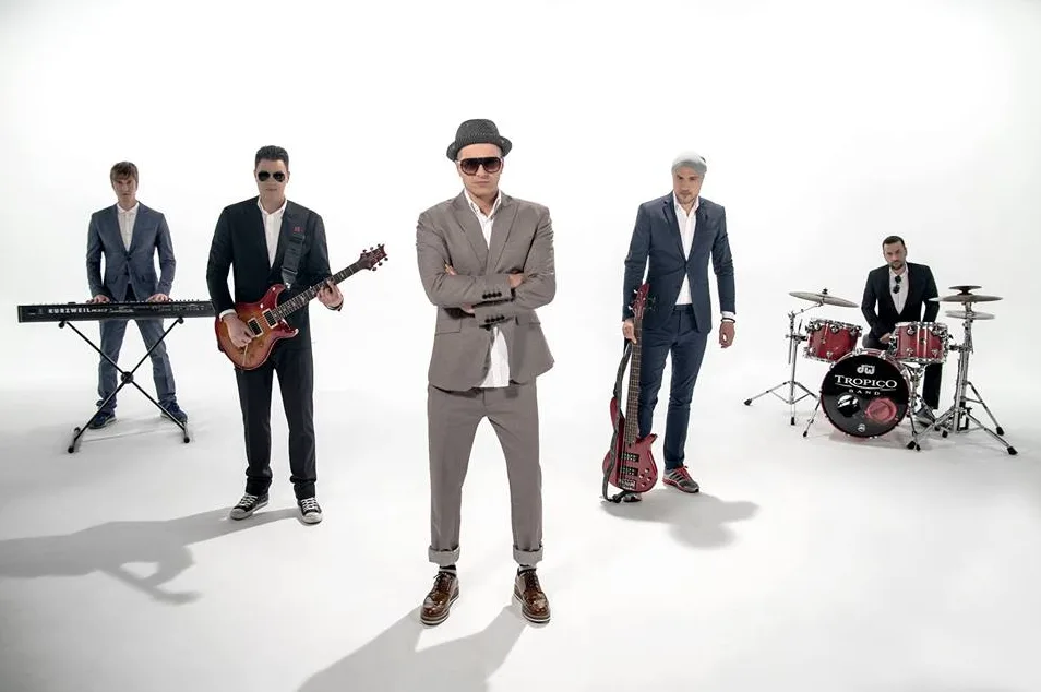 Pet članani bend "Tropicо Band" pozira za fotografiju u studiju. U prvom planu, u centru, nalazi se pjevač sa šeširom i naočalama, obučen u odijelo. S lijeva na desno, stoje klavijaturista, gitarista, basista i bubnjar za bubnjevima. Bend je obučen u elegantna odijela.