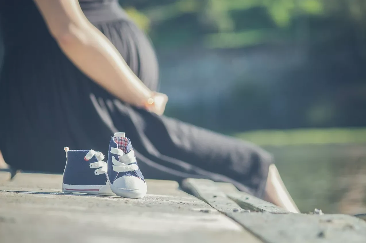 Trudnica u crnoj haljini sjedi pored malih plavih cipela namijenjenih bebi.