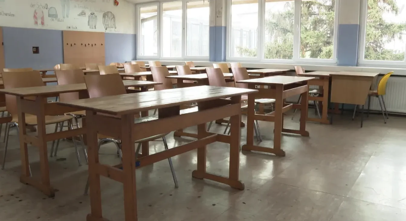 Unutrašnjost prazne učionice sa redovima drvenih klupa i stolica, osvijetljene velikim prozorima kroz koje se vidi drveće.