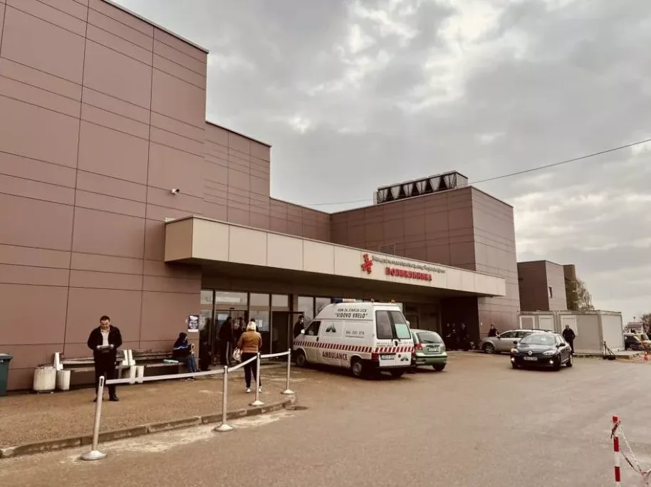 Ulaz u savremenu bolnicu sa parkiranim vozilima ispred, uključujući kombi hitne pomoći.